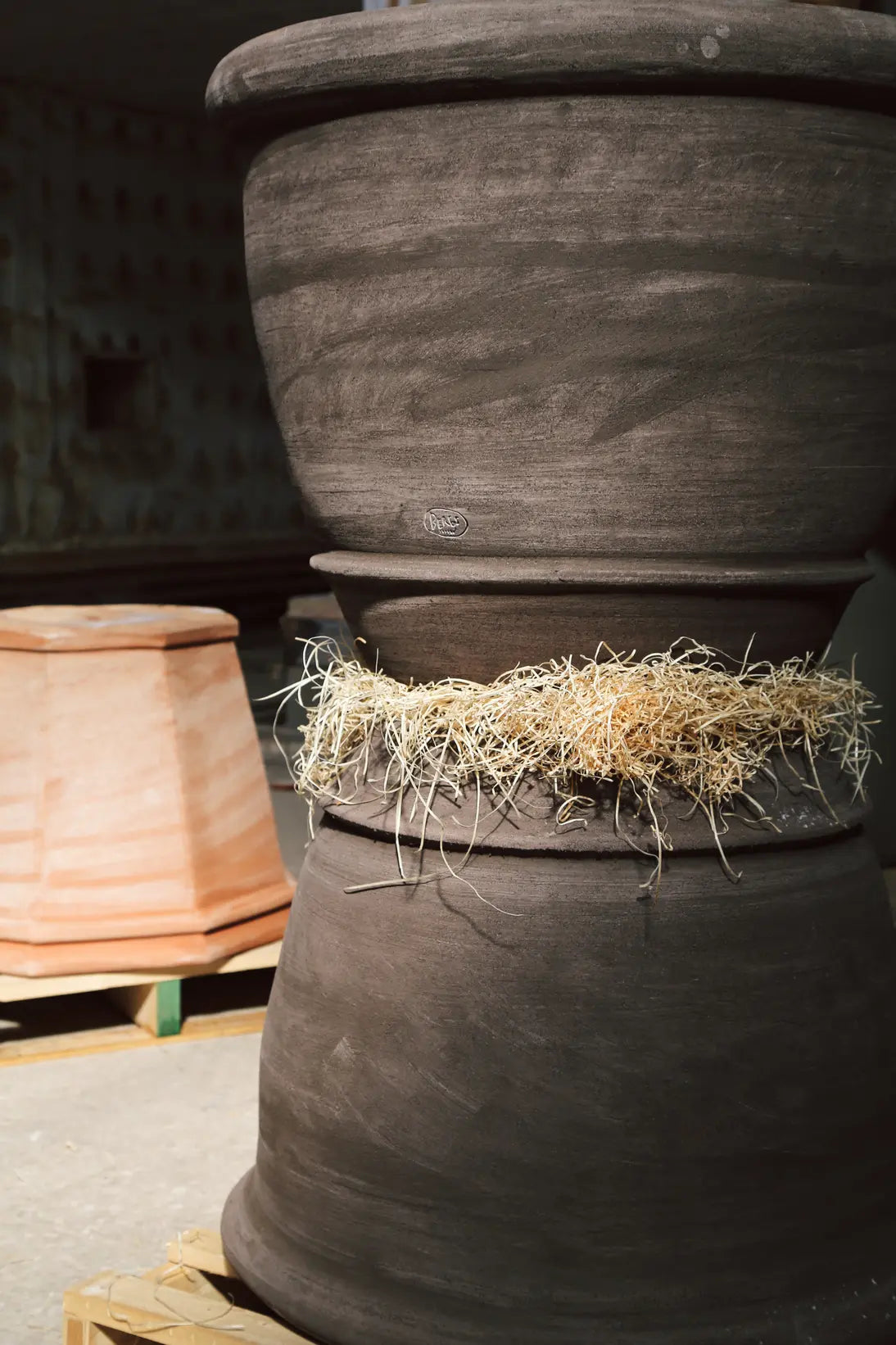 2 large gray clay pots stacked on top of one another, with a terracotta pot resting upside down on a pallet in the background.